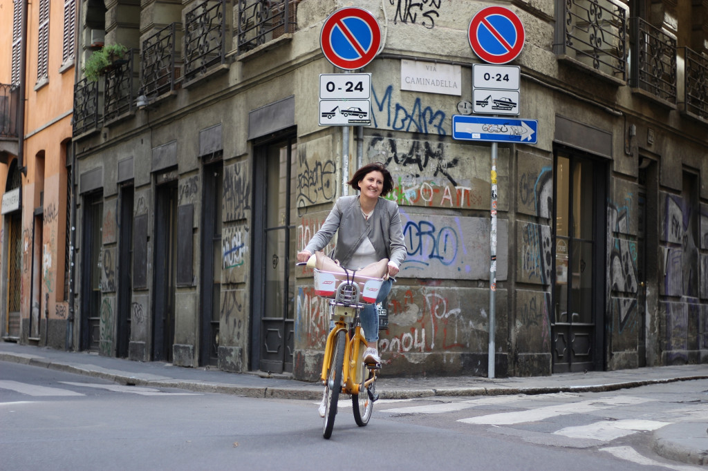 In der Bildmitte fährt eine Frau auf einem gelben Fahrrad auf die Kamera zu. Im Hintergrund eine mit Graffiti verzierte Häuserwand. Radfahren in Mailand.