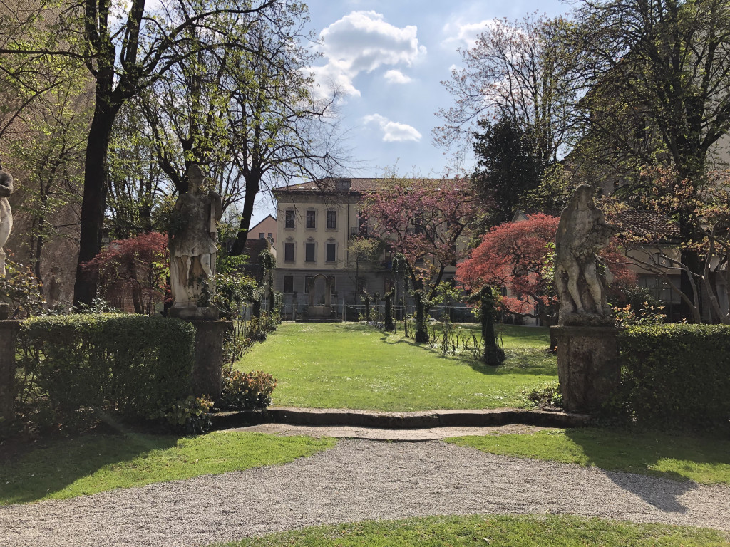 Blick in den Garten desCasa degli Atellani: Grüner Rasen, im linken Bildrand eine Hecke, dahinter Bäume. Auch auf dem Rasen stehen niedrige Bäume. Im Bildhintergrund ist ein langgezogenes Haus zu sehen.