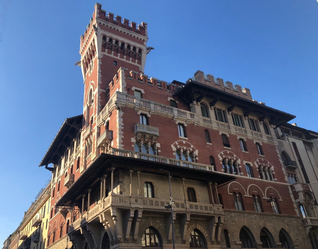 In unmittelbarer Nähe zu Sant'Ambrogio befindet sich das Castello Corvo. Hier bildfüllend vor strahlend blauem Himmel. Ein rot-braune Fassade mit Türmchen, Vorsrprüngen, Balkönchen und Zinnen 