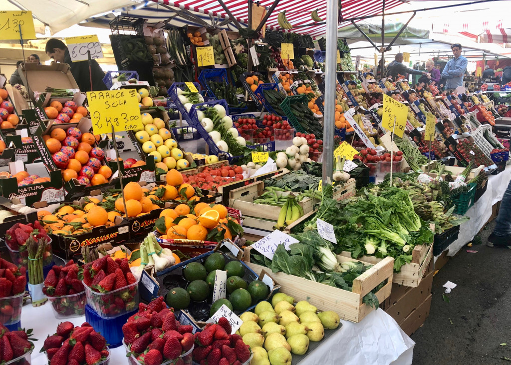 Mailand-Tipps: Ein Tag rund um Sant‘Ambrogio. Bildfüllend ist ein Obst- und Gemüsestand zu sehen. Feil geboten werden im Vordergrund Erdbeeren, Birnen, Avocados, Orangen. Alle in Kisten angeboten. Im Hintergrund zwei Männer, die sich unterhalten und weitere Markstände. 