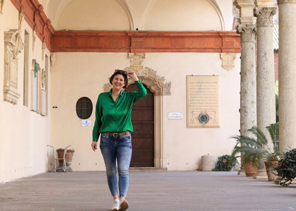 Frau im Bogengang des Cortile della Canonica de Sant’Ambrogio Eine Frau in Jeans und knallgrüner Bluse steht in einem hellen Bogengang der mit Backsteinbögen. In der rechten Bildhälfte stehen Säulen mit korinthischen Kapitellen und Pulvino, dazwischen ein paar niedrige Palmen im Cortile della Canonica de Sant’Ambrogio