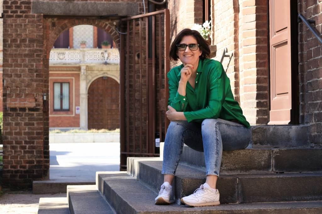 Eine Frau mit Sonnenbrille und dunklen halblangen Haaren sitzt in knallgrüner Bluse und Jeans auf Stufen vor einer Holztüre- Im Hintergrund eine offenes Tor das den Blick frei gibt auf das dahinterliegende Haus mit oben abgerundeter Holztür.