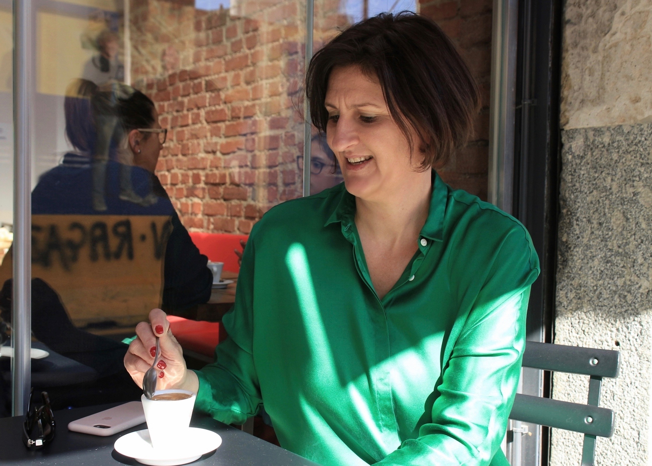 Wie sieht ein italienisches Frühstück aus? In der rechten Bildhälfte sitzt eine Frau in grüner Bluse. Vor ihr auf dem Tisch eine Espressotasse. Die Frau hält in der rechten Hand einen Kaffeelöffel und taucht ihn in den Espresso. Neben der Tasse liegt ein Smartphone.