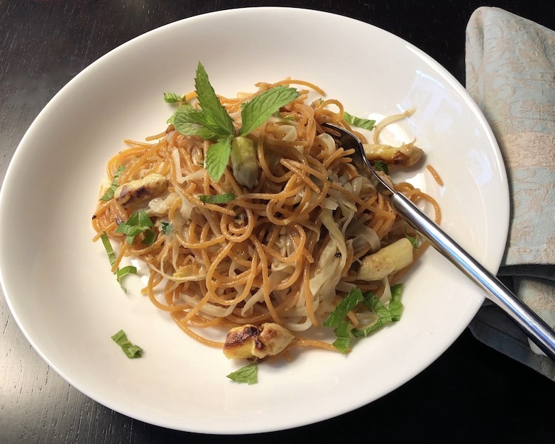 Ein beigefarbener Pastateller mit rötlichen Spaghetti aus roten Linsen, weißem Spargel und Minzblättchen. In der Pasta steckt eine Gabel, die rechts aus dem Teller reicht. 