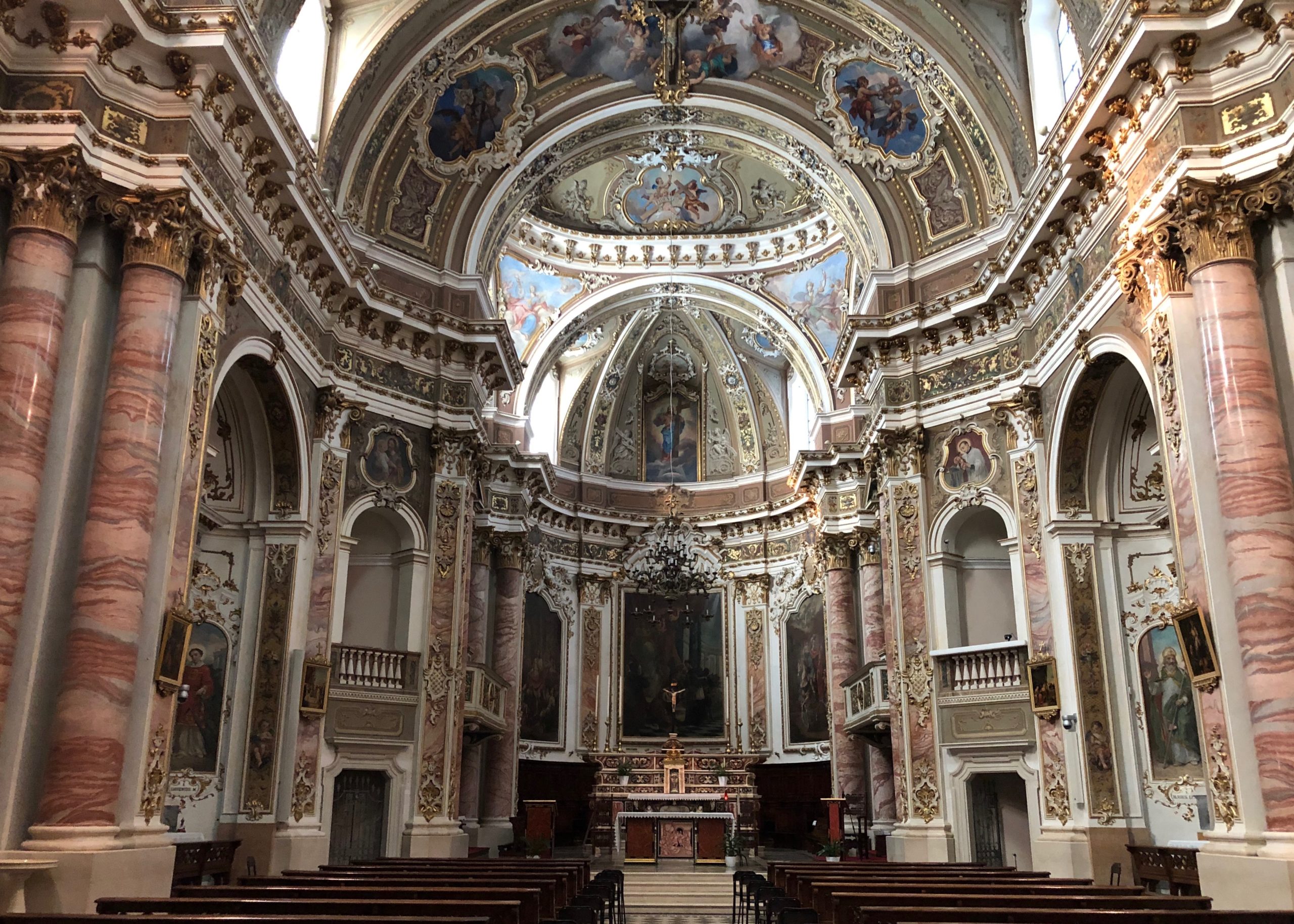 Innenansicht der barocken Kirche San Martino mit Blick in den reich dekorierten Altarraum, schlicht dunkelbraune Holzkirchenbänke und Säulen aus rosa Marmor und Akantuskapitellen an den Seiten