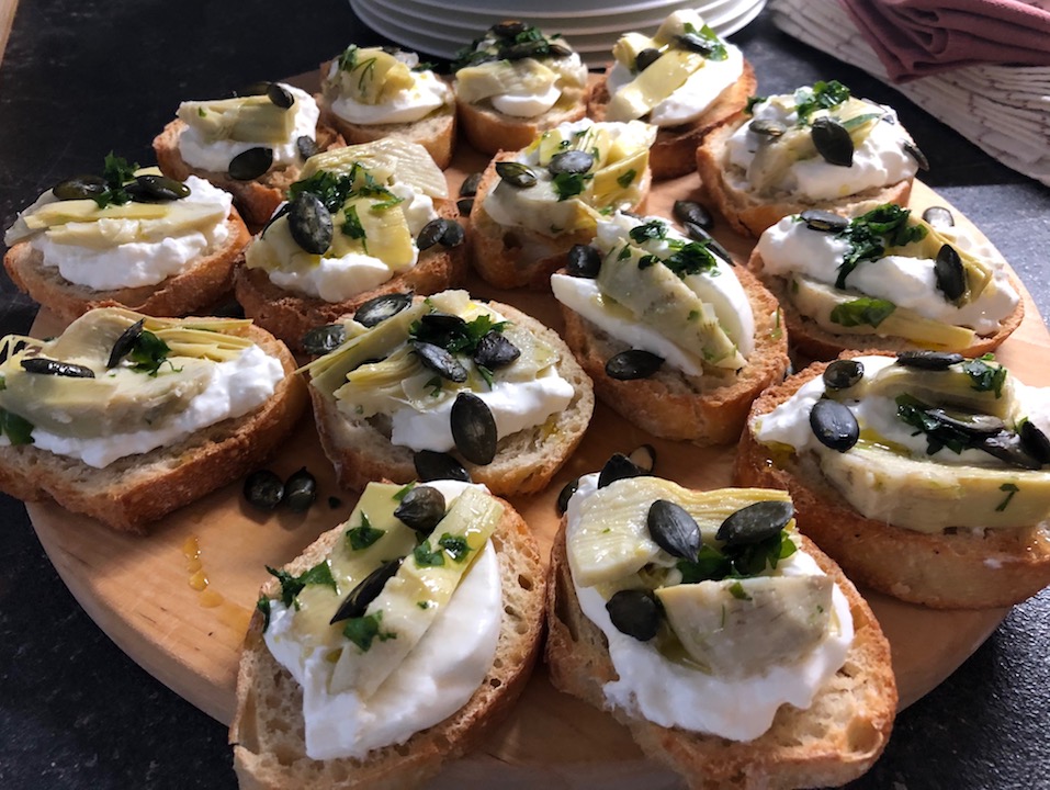 Crostini mit Mozzarella und Artischocken Bildfüllende liegt auf dunklem Untergrund ein rundes Holzbrett. Darauf sind Crostini, kleine geröstete Brotscheiben, verteilt auf denen eine Scheibe Büffelmozzarella und geschnittene eingelegte Artischocken liegen. Kürbiskerne und Olivenöl sind darüber geträufelt.