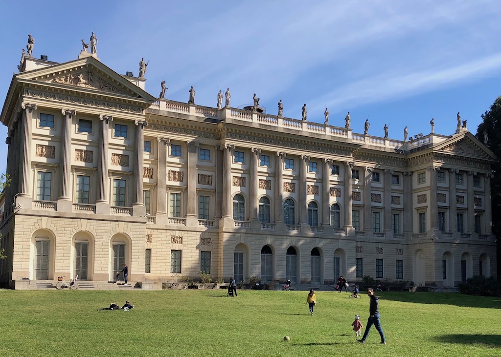 Blick auf die Villa Reale vom Park aus, ein Geheimtipp für den nächsten Mailand-Besuch Geheimtipp in Mailand: Die Villa Reale, von der im Hintergrund die Gartenseite der Gebäudefassade im Neoklassizismus zu sehen ist. Im Vordergrund eine Rasenfläche auf der ein paar spielende Kinder zu sehen sind. Einer meiner Geheimtipps für den nächsten Mailand-Besuch