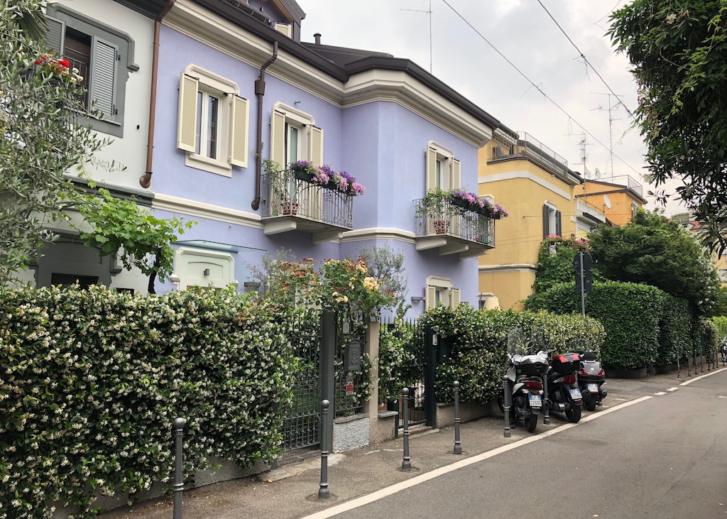 Kolorierte Häuserfassade im Villagio Operaio di Via Lincoln in Mailand Eine Häuserfront, zweistöckig mit Fensterläden, kleinen Balkonen und Blumenschmuck. Gestrichen sind die Häuser in lila, gelb und orange. Davor sind Gartenzäune von Büschen umrankt. Im Vordergrund eine Straße. Einer meiner Geheimtipps für den nächsten Mailand-Besuch