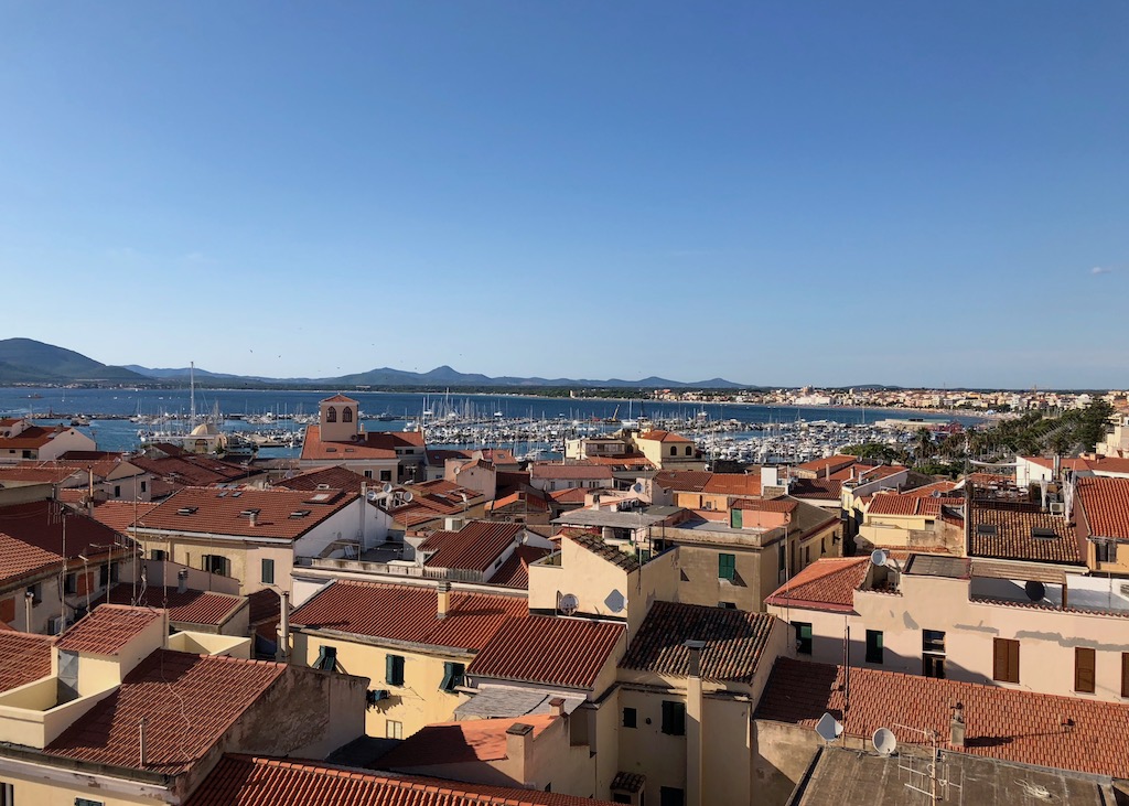 Blick über rote flache Ziegeldächer in Alghero bei klarem Himmel. Im Hintergrund ist das blaue Meer mit Segelschiffen zu sehen. 