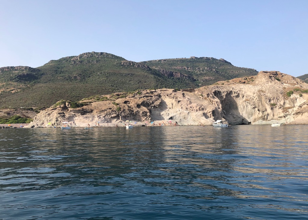 Ein Sommertag in der Nähe von Bosa auf Sardinien. Das Foto ist vom Wasser aus aufgenommen. Im Bildvordergrund stilles tiefblaues ruhiges Wasser. Dahinter eine helle steile Steinküste und noch eine Ebene dahinter begrünte Berge. Darüber blauer Himmel. 