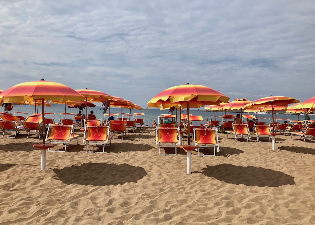 Eine Strandbad in Bosa Marritima. In mehreren Reihen stehen orangefarbene Sonnenschirme, darunter jeweils zwei orangefarbene Liegestühle. Im Hintergrund ist das Meer. 