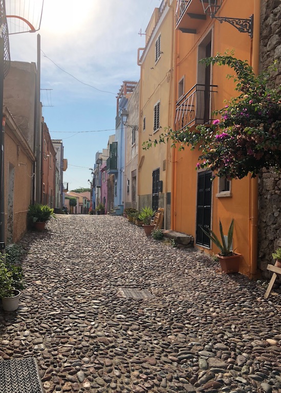 Zentral im Bild die für Bosa so typischen gepflasterte schmale Gasse. Links und rechts davon kolorierte Häuser.