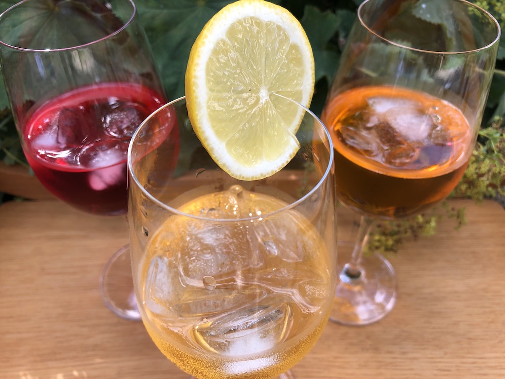 Aperitifs ohne Umdrehungen. Auf einem hellen Brett stehen drei langstielige Gläser, die halb voll sind als Dreieck angeordnet. Das Glas im Vordergrund ist mit einem gelben Getränk (Martini Floreale) und mehreren Eiswürfel gefüllt. Am Glasrand steckt eine Scheibe Zitrone. Das Glas links davon im Hintergrund ist mit einem rotfarbenen Getränk (Martini Vibrante) und Eiswürfeln gefüllt, das rechte mit einem orangefarbenen Getränk (Crodino) und mehreren Eiswürfeln.