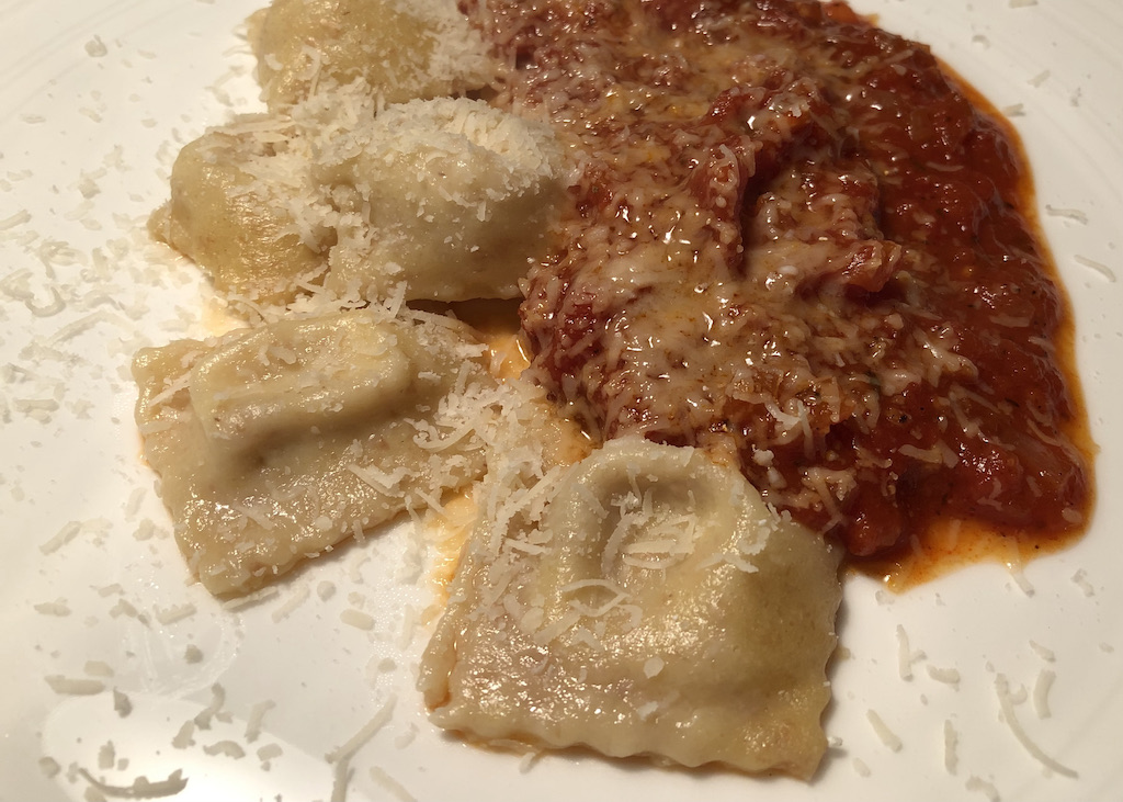 Auf einem beigefarbenen Teller liegen eine Tomatensoße und darauf kleine quadratische Ravioli, die mit geriebenem Parmesan bestreut sind. Selbst gemachte Ravioli mit einer Mozzarella-Zimt-Füllung