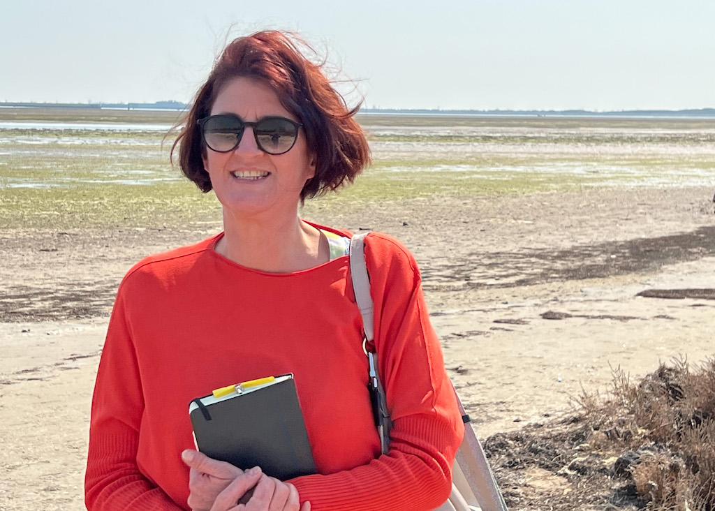 Slow Travel in Venedig: Ein geführter Ausflug auf die Insel Sant‘Erasmo Im Vordergrund eine Frau mit dunklen halblangen Haaren und Sonnenbrille in orangefarbenem Pulli. Über der Schulter hängt eine helle Ledertasche. Im Hintergrund ist eine trockene Salzwiese zu sehen.