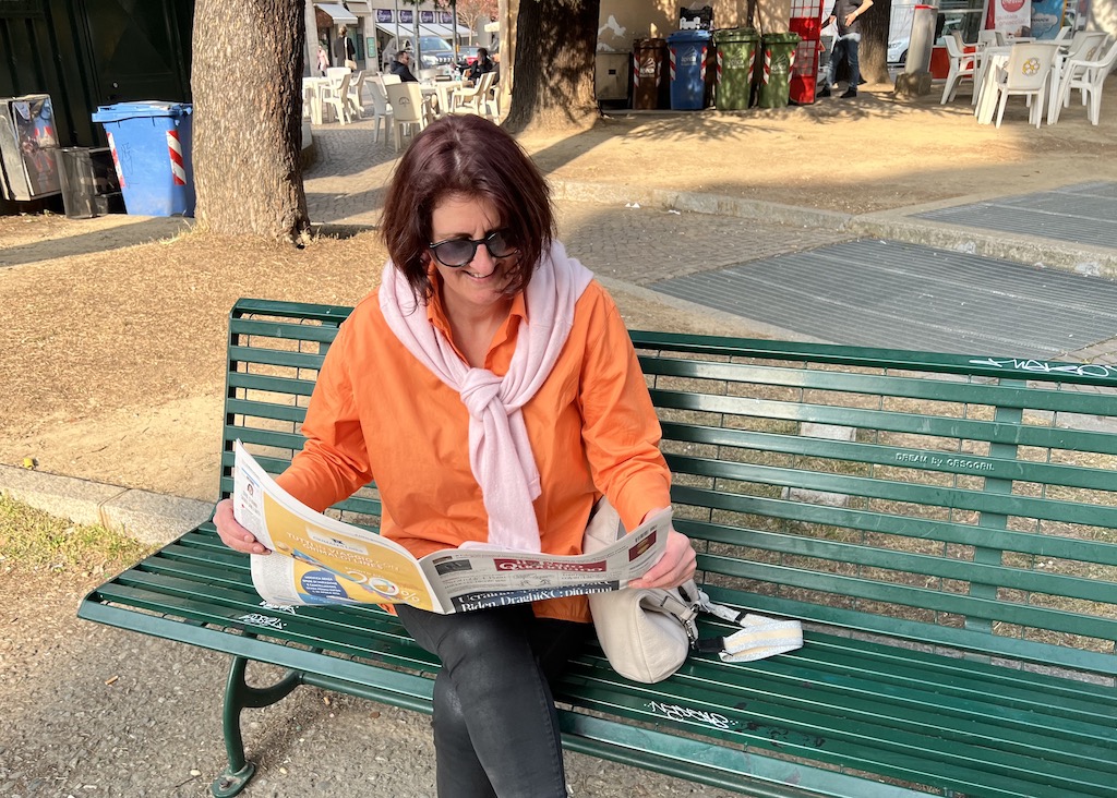 Auf einer grünen Bank sitzt eine Frau mit Sonnenbrille und dunklen Haaren in einer orangefarbener weiten Bluse und schwarzer Hose. In den Händen hält sie die aufgeschlagene italienische Zeitung il Fatto Quotidiano, die sie interessiert liest. 