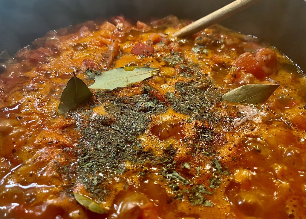 Topf mit Tomatensoße In einem dunklen Gusseisentopf köchelt eine Tomatensoße mit Lorbeerblättern, getrocknetem Oregano, Muskat, Pfeffer, Salz