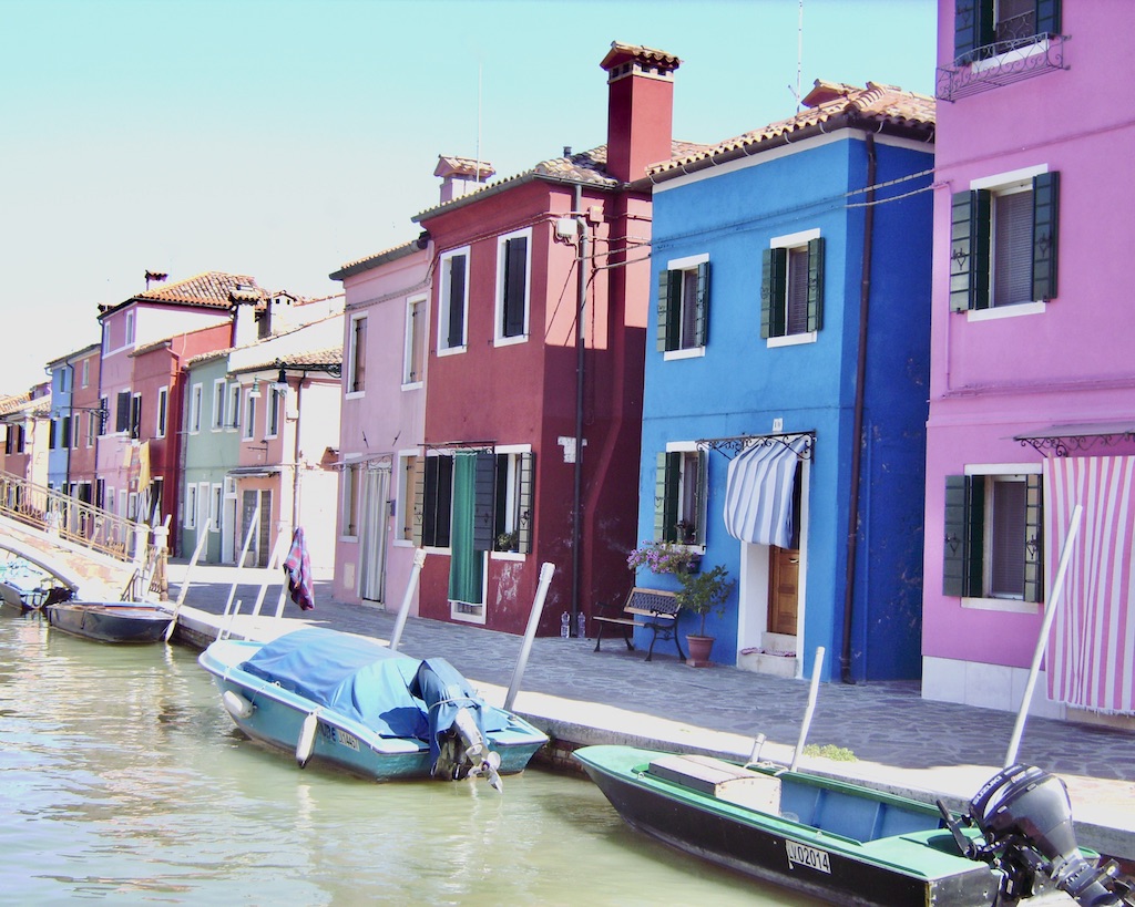 Eng nebeneinander stehende farbige Häuser (pink, blau.rostrot, rosa, mint) Davor ein Kanal mit Booten am Ufer auf Burano in der Lagune Venedigs