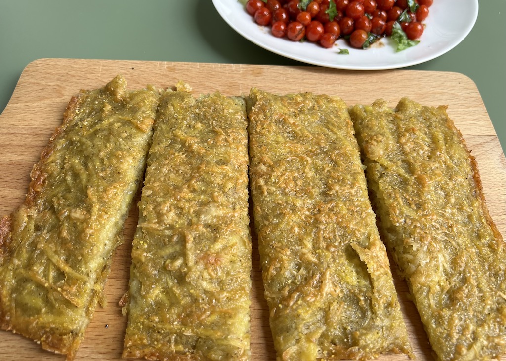 Auf einem Holzbrett liegen in vier schmale lange Stücke geschnittene knuspriger Parmesan-Kartoffelfladen. Im Hintergrund liegen auf einem beigen Teller in Olivenöl kurz gedünstete Minitomaten mit Basilikum 