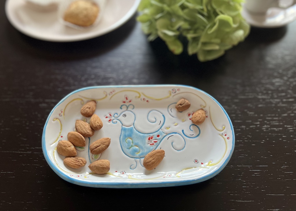 Ovale Keramikschale mit einigen Mandeln darauf. Im Zentrum ein Hahn - typisches Motiv für Sardinien- Im Hintergund ein Teller darauf Papassinos, daneben eine grüne Blüte und eine Espressotasse 