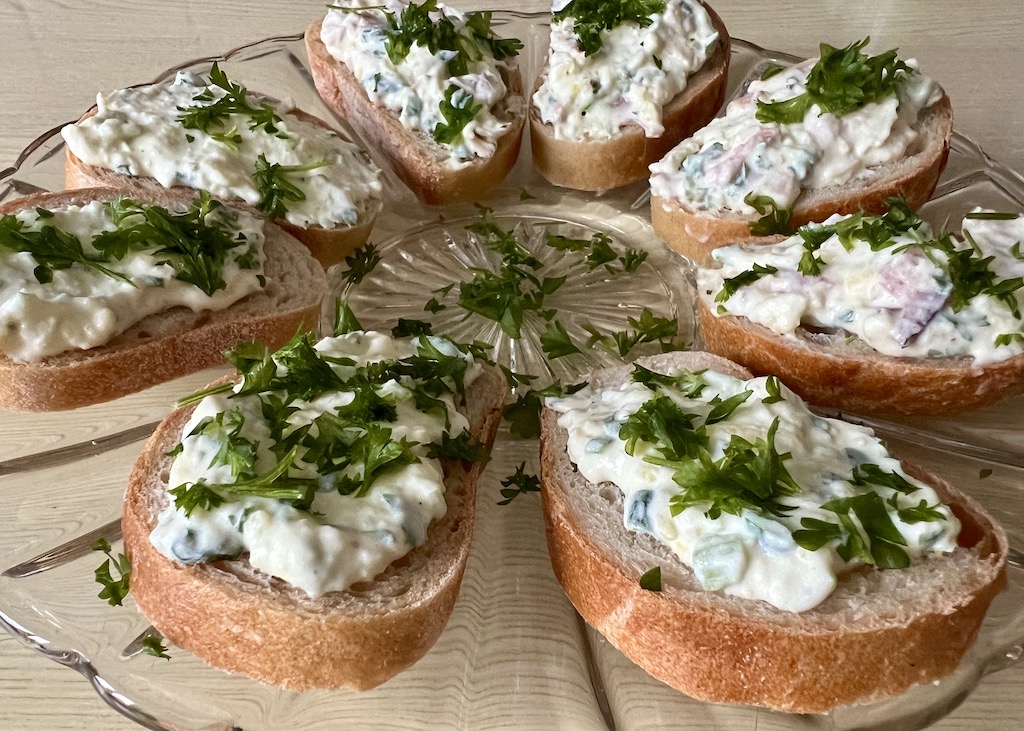 Crostini mit einer leckeren Kartoffelcreme: Auf einem Glasteller liegen mehrere Baguettescheiben, die mit einer Kartoffel-Schmandcreme bestrichen sind. Dekoriert sind sie mit Petersilieblättchen