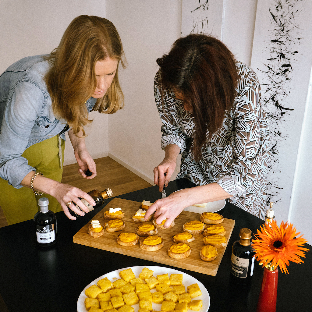 Vorbereitungen fürs aperitivo-Buffet. Vanessa Wanhoff von balsAmici (links) und Ulrike Schmid von pasticcio.eu (rechts) verfeinern Crostini mit Kürbiscreme mit Brie und Aceto Balsamico. Im Vordergrund ein beiger Teller mit Polentawürfeln.