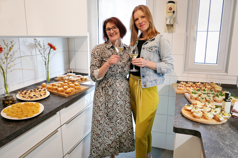 Vanessa Wanhoff BalsAmici Ulrike Schmid pasticcio.eu Im Bildzentrum stehen Vanessa Wanhoff von BalsAmici (r) und Ulrike Schmid von pasticcio.eu (l) lachend mit einem Glas Weißwein in der Hand. Links und rechts von ihnen stehen auf einer Küchenablage verschiedene Häppchen, die noch mit Aceto Balsamico verfeinert werden.