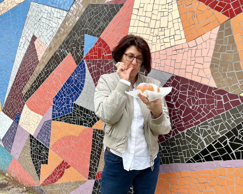 Vor einer farbigen Mosaikwand mit geometrischen Mustern steht eine Frau. In einer Hand hält sie einen maritozzo auf einer Serviette. Den Daumen der anderen Hand befreit sie gerade von geschlagener Sahne. 