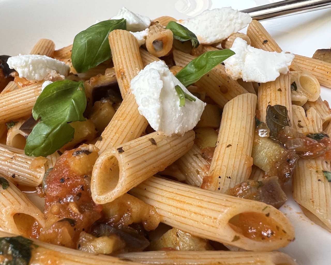 Pasta alla Norma: Auf einem beigen Teller liegen durchmischt Penne Rigate, gebratenene Tomaten-und Auberginenstücke, darüber Ricotta und Basilikumblättchen.