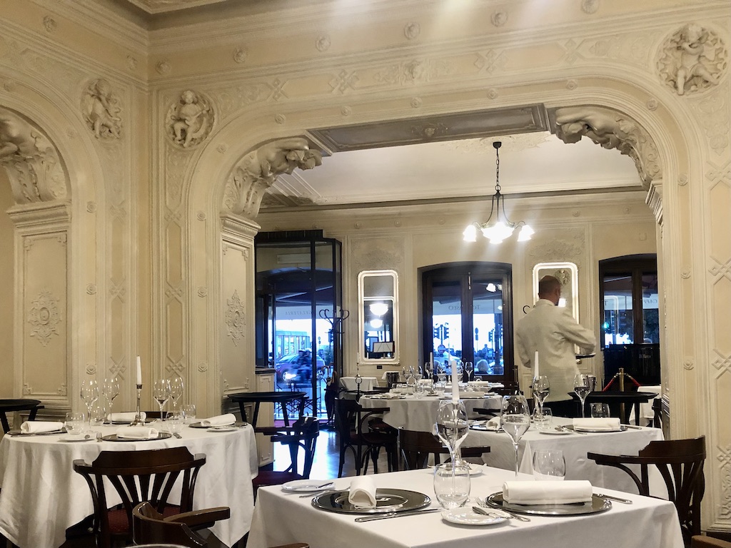 Italiens historische Cafes: Innenansicht des Caffé Tommasseo Triest. Quadratische Tische mit weißen gestärkten Tischdecken. Darauf silberne Platzteller, Stoffservieten und Besteck. An den Wänden Stuckverzierungen