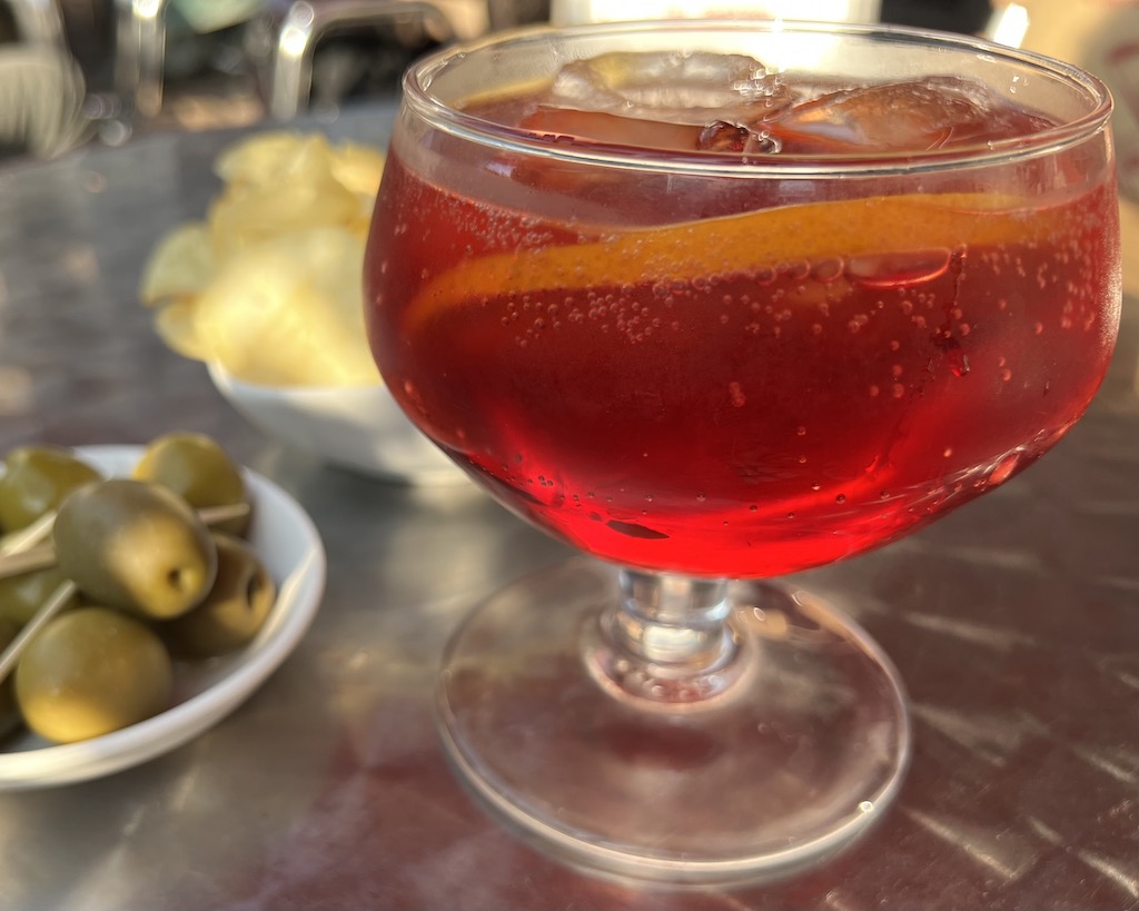 Negroni Sbagliato Bar Basso Milano Auf einem kleinen Tisch steht Glas, gefühlt mit einem Negroni Sbagliato. Daneben steht ein Schälchen Chips und ein Tellerchen mit grünen Oliven.
